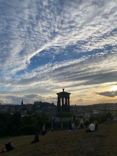 Ein Monument während des Sonnenuntergangs, davor eine Wiese mit Menschen, im Hintergund Blick auf die Stadt Edinburgh