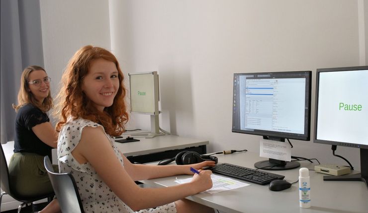 Eine Versuchsleiterin und eine Versuchsperson sitzen in einem Computerlabor und lächeln in die Kamera. Auf dem Monitor ist das Wort „Pause“ zu sehen.