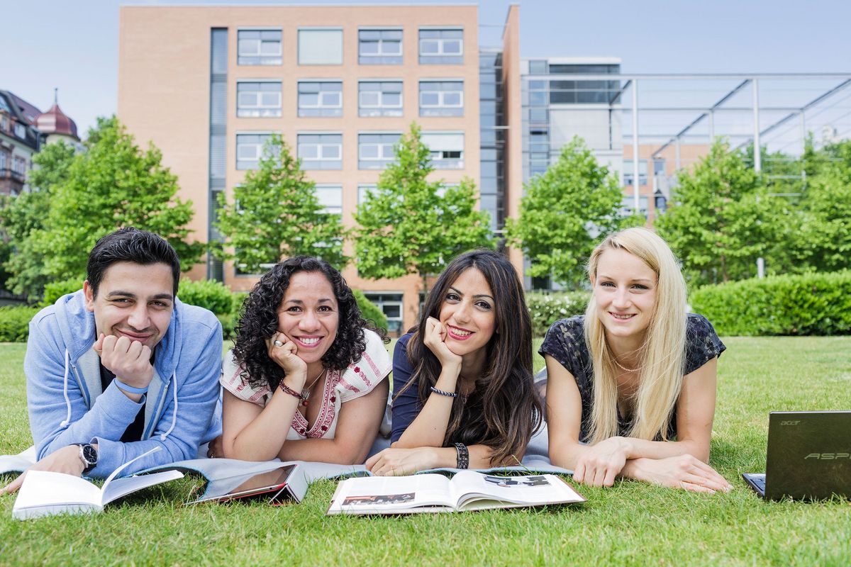 zur Vergrößerungsansicht des Bildes: Eine Gruppe internationaler Studierender liegt vor der Universität Leipzig in der Sonne. Die Studierenden blicken von ihren Lehrmaterialien auf und schauen glücklich in die Kamera.