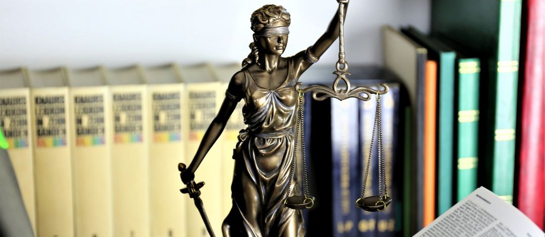 Farbfoto: eine kleine bronzefarbene Statue der Justizia mit erhobenem Schwert, verbundenen Augen und Waagschale steht auf einem Tisch. Im Vordergrund liegt ein aufgeschlagenes Buch mit Gesetzestexten. Im Hintergrund stehen Bücher in einem Regal.