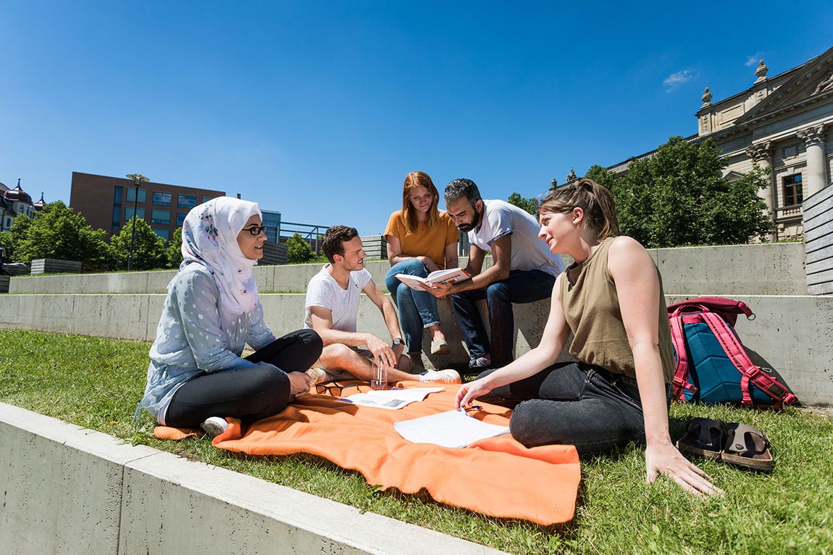 zur Vergrößerungsansicht des Bildes: Internationale Studierende sitzen auf einer Picknickdecke in der Sonne vor dem Campus Beethovenstraße 