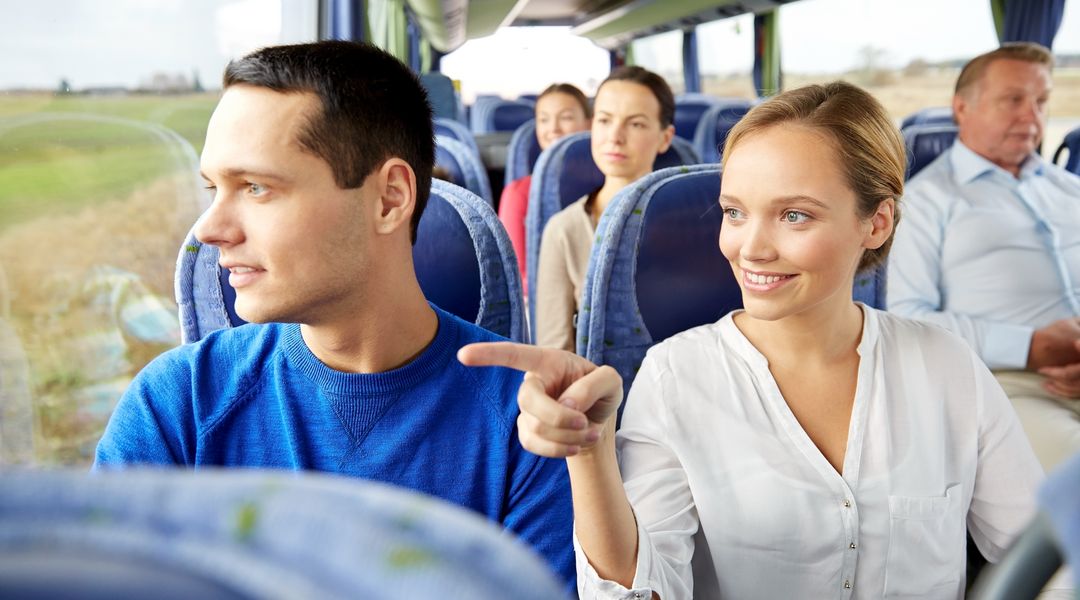 Personen sitzen in einem Bus. Eine Frau zeigt ihrem Sitznachbarn etwas draußen.