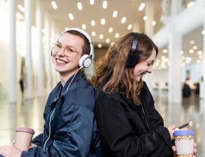 Zwei Studentinen sitzen Rücken an Rücken im Augusteum und haben Kopfhörer auf. Die linke Studentin blickt lachend in die Kamera. Die rechte Studentin schaut lächenlnd auf ihr Smartphone, welches sie in der Hand hält.