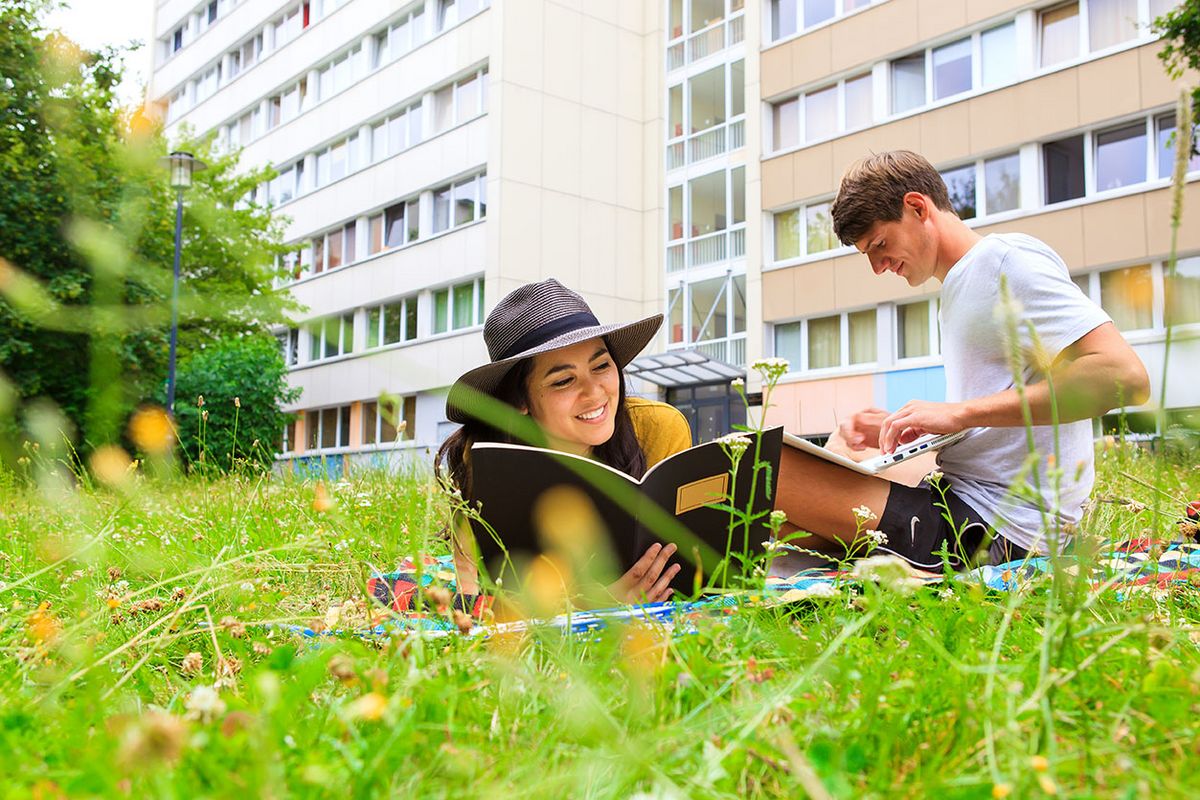 enlarge the image: Zwei Studierende der Universität Leipzig liegen vor dem Studentenwohnheim im Gras. Sie genießen die Sonne und sind in Lernunterlagen vertieft.