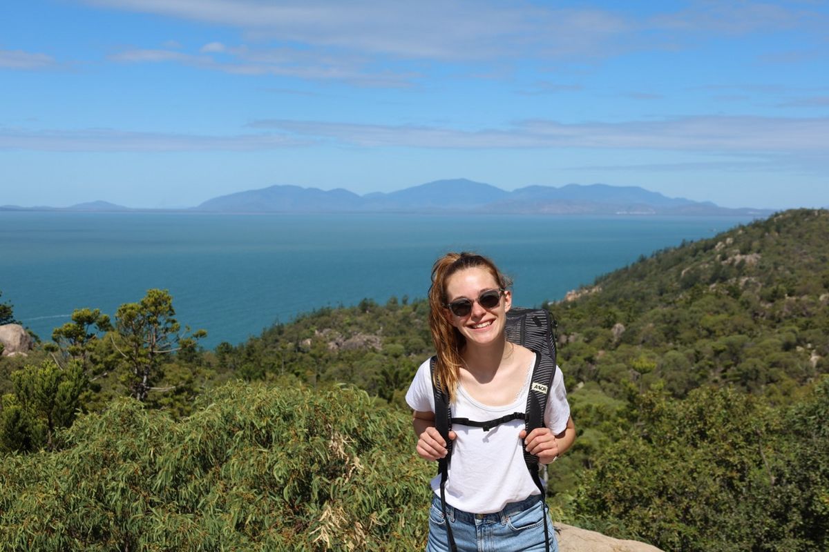 zur Vergrößerungsansicht des Bildes: Studentin Estelle in Australien im Hintergrund Meer und Berge, sie steht inmitten von Grün 