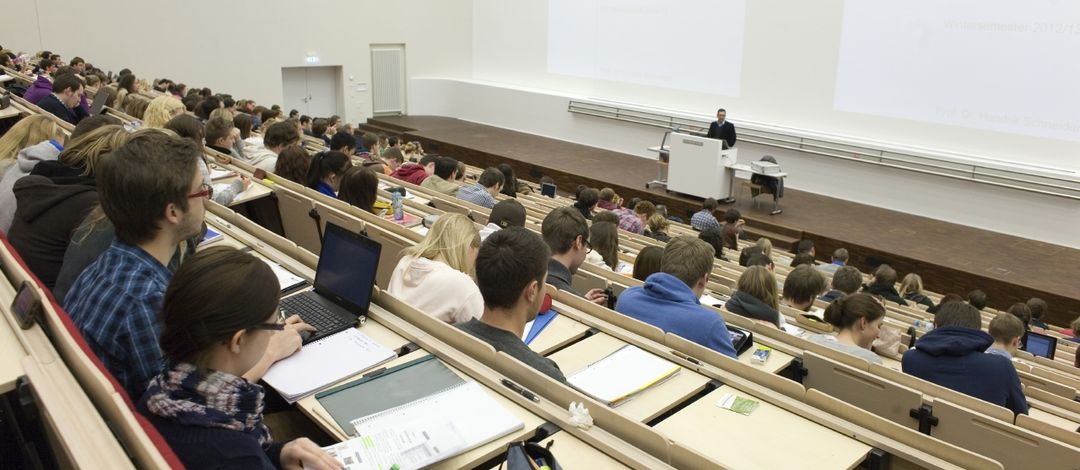 Foto: Vorlesung im vollbesetzten Audimax, am Rednerpult steht ein Professor