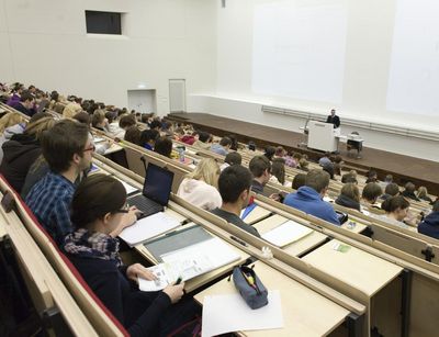 Foto: Vorlesung im vollbesetzten Audimax, am Rednerpult steht ein Professor