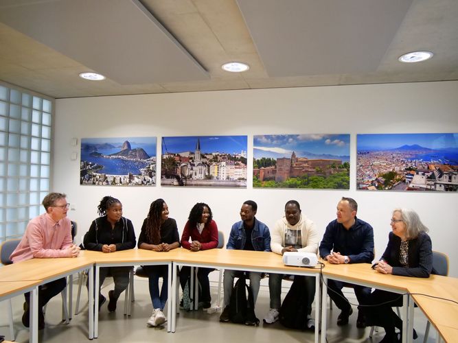 Gruppenbild der fünf mosambikanischen Austauschstudierenden im Gespräch mit ihren drei Leipziger Betreuer:innen im Seminarraum des Geisteswissenschaftlichen Zentrums der Universität Leipzig. Die Gruppe sitzt an Holztischen. An der weißen Wand im Hintergrund hängen vier großformatige Fotos mit Landschafts- und Stadtansichten.