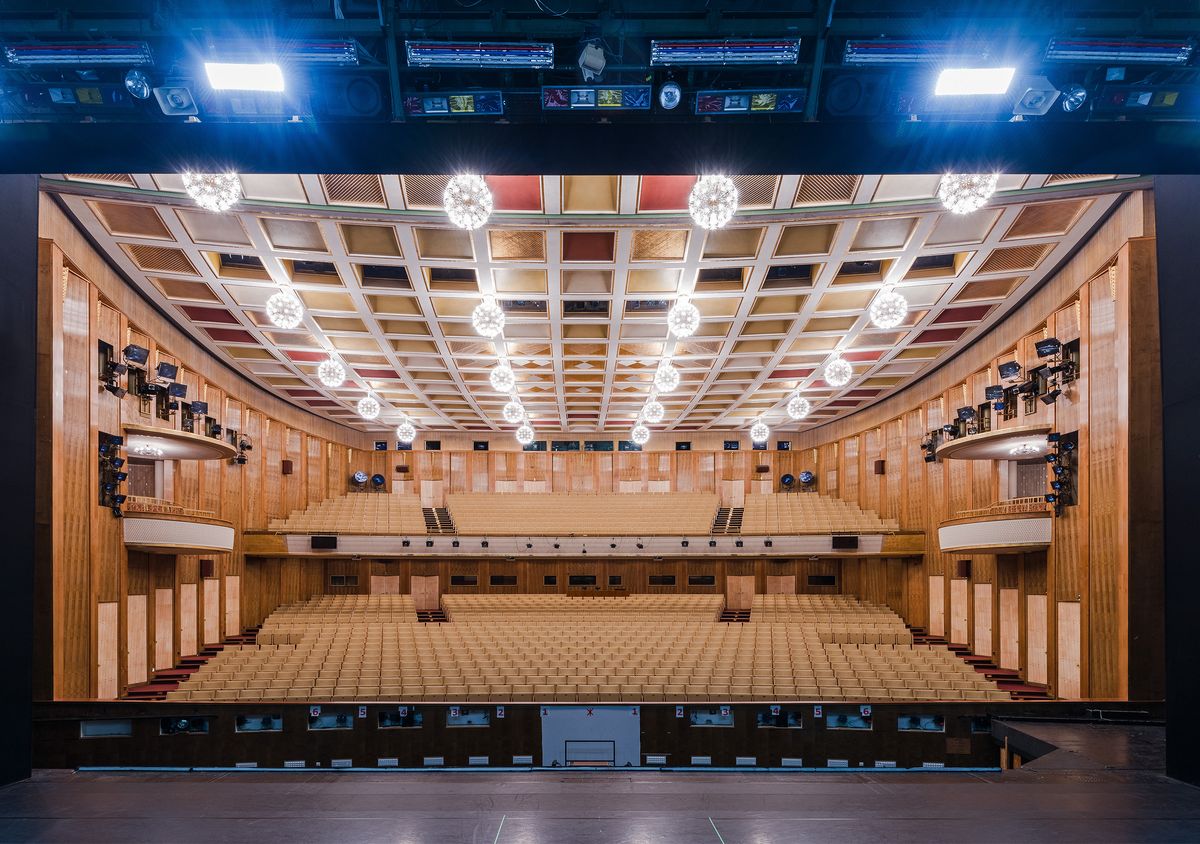 Das Foto zeit einen großen Saal von der Bühne aus betrachtet. Die Wänge wirken hölzern verkleidet, an der Decke leuchten viele, helle Lampen. Der Saal gehört zur Oper Leipzig, man sieht ihn aus der Perspektive von Opernsingenden.