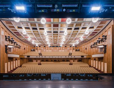 Das Foto zeit einen großen Saal von der Bühne aus betrachtet. Die Wänge wirken hölzern verkleidet, an der Decke leuchten viele, helle Lampen. Der Saal gehört zur Oper Leipzig, man sieht ihn aus der Perspektive von Opernsingenden.