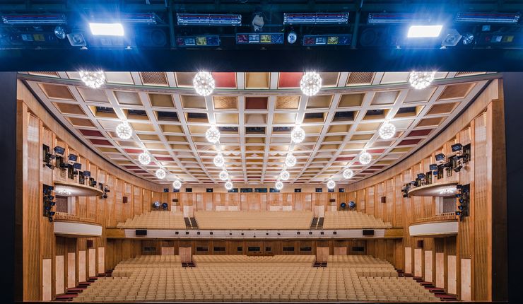 Das Foto zeit einen großen Saal von der Bühne aus betrachtet. Die Wänge wirken hölzern verkleidet, an der Decke leuchten viele, helle Lampen. Der Saal gehört zur Oper Leipzig, man sieht ihn aus der Perspektive von Opernsingenden.