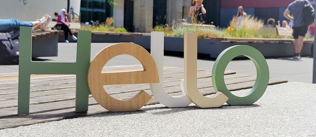 Detailaufnahme: Der Schriftzug "Hello" aus bunten Holzbuchstaben steht auf einer Bank. Im Hintergrund ist der Innenhof vom Campus Augustusplatz mit viel Grün zu erkennen.