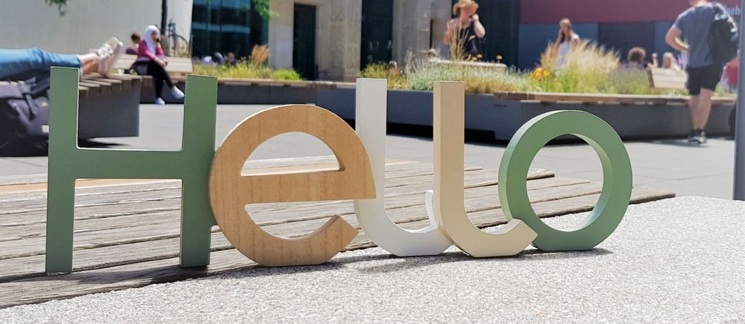 Detailaufnahme: Der Schriftzug "Hello" aus bunten Holzbuchstaben steht auf einer Bank. Im Hintergrund ist der Innenhof vom Campus Augustusplatz mit viel Grün zu erkennen.