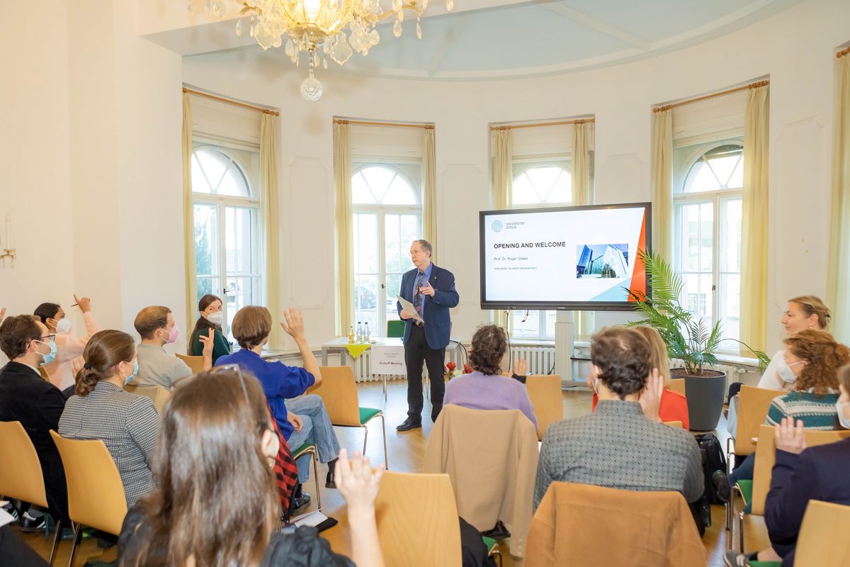 zur Vergrößerungsansicht des Bildes: Ein heller Raum, in dem viele Menschen auf Stühlen sitzen. Vor ihnen steht Prorektor Prof. Glaeser und begrüßt sie zur Auftaktveranstaltung.
