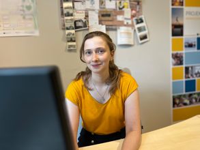 Frau sitzt hinter einem Computerbildschirm am Arbeitsplatz und lächelt in die Kamera