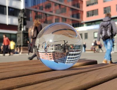 Farbfoto: Eine Glaskugel liegt auf einer Bank und in Ihr spiegeln sich Menschen und Gebäude aus dem Hintergrund