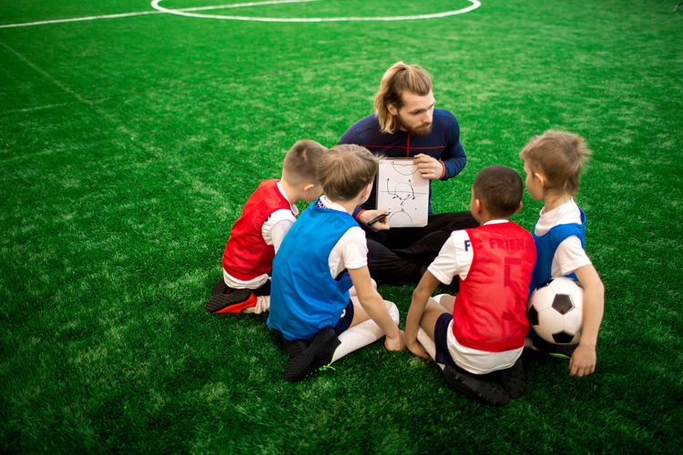 Zu sehen ist ein Trainer auf einem Fußballfeld, der Kindern an einer Taktiftafel Spielregeln erklärt.