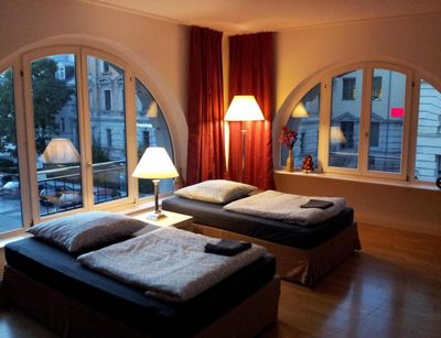 Farbfoto: Innenansicht eines Wohnraums mit zwei Betten, bodentiefen Fenstern und einer Stehlampe