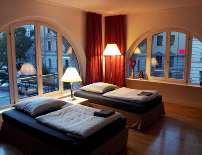 Colour photo: Interior view of a living room with two beds, floor-to-ceiling windows and a floor lamp