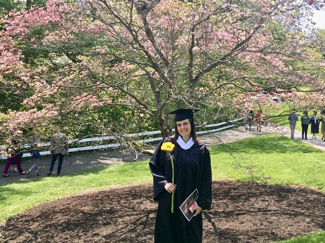 Die Studierende steht lächelnd in ihrer schwarzen Graduationsrobe vor einem blühenden Baum und hält eine Sonnenblume sowie ihr Zeugnis in den Händen.