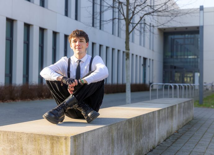 Noah Dejanovic sitzt im Außenbereich des Campus Jahnallee auf einem Stein und schaut lächelnd in die Kamera.