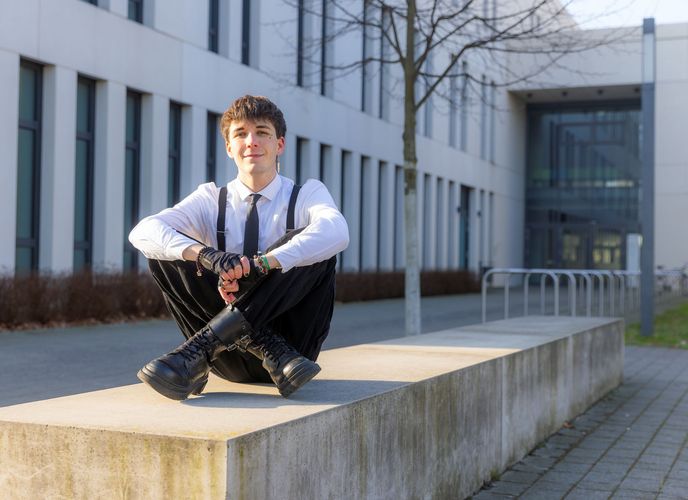 Noah Dejanovic sitzt im Außenbereich des Campus Jahnallee auf einem Stein und schaut lächelnd in die Kamera.