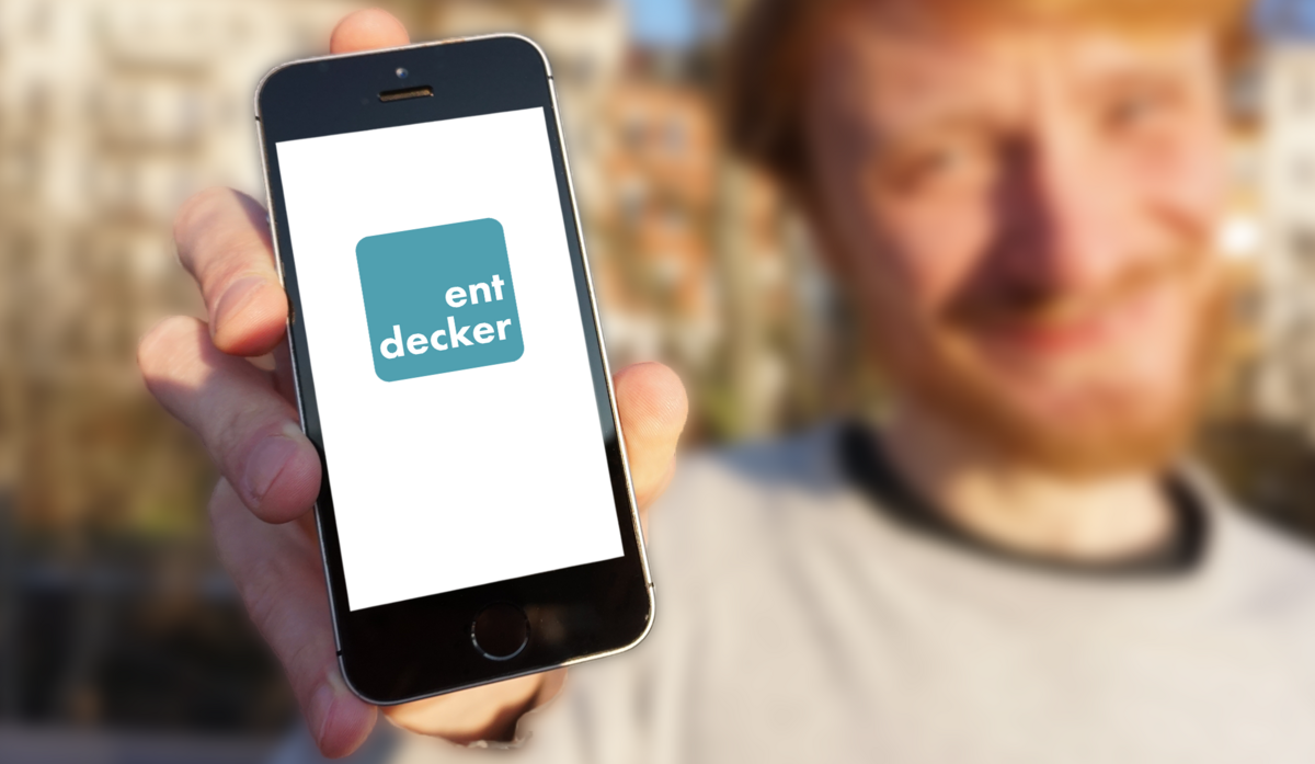 enlarge the image: Auf dem Bild sieht man im Hintergrund einen jungen Mann, der in seiner Hand ein Smartphone hält. Das Smartphone zeigt er in die Kamera. Auf dem Smartphone ist ein Logo mit dem Schriftzug "entdecker" zu sehen.