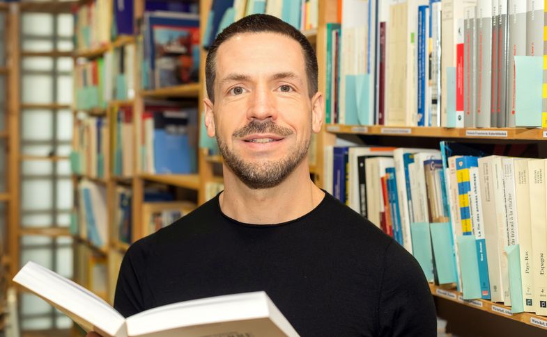 Prof. Klaus Grübel steht mit einem offenen Buch in der Bibliothek
