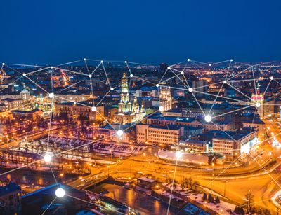Farbfoto: Vogelperspektive auf eine erleuchtete Stadt in der Nacht. Über dem Bild liegen grafische Elemente 