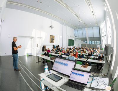 A monitor and a laptop showing recording traces with a lecturer nearby and audience sitting in the background 
