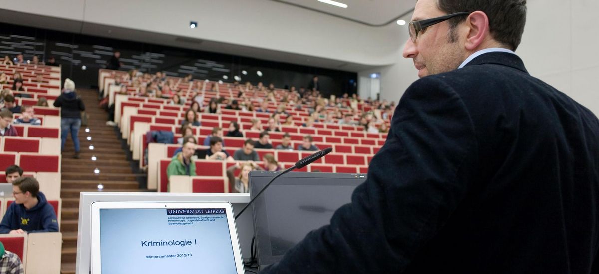 Foto: ein Mann steht am Rednerpult im voll bestetztem Hörsaal und spricht zu den Studierenden während er auch auf einen Laptop blickt.