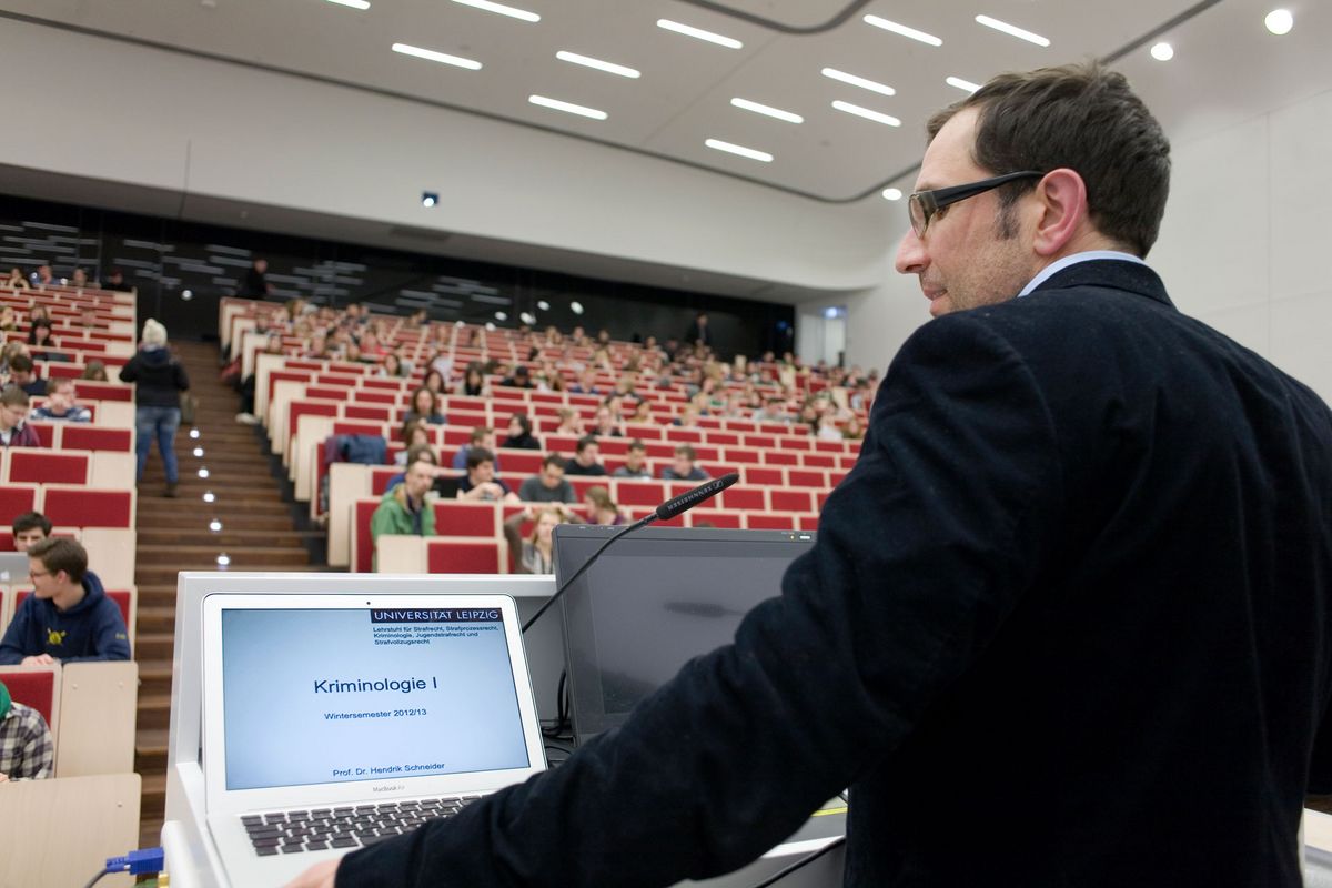 zur Vergrößerungsansicht des Bildes: Foto: ein Mann steht am Rednerpult im voll bestetztem Hörsaal und spricht zu den Studierenden während er auch auf einen Laptop blickt.