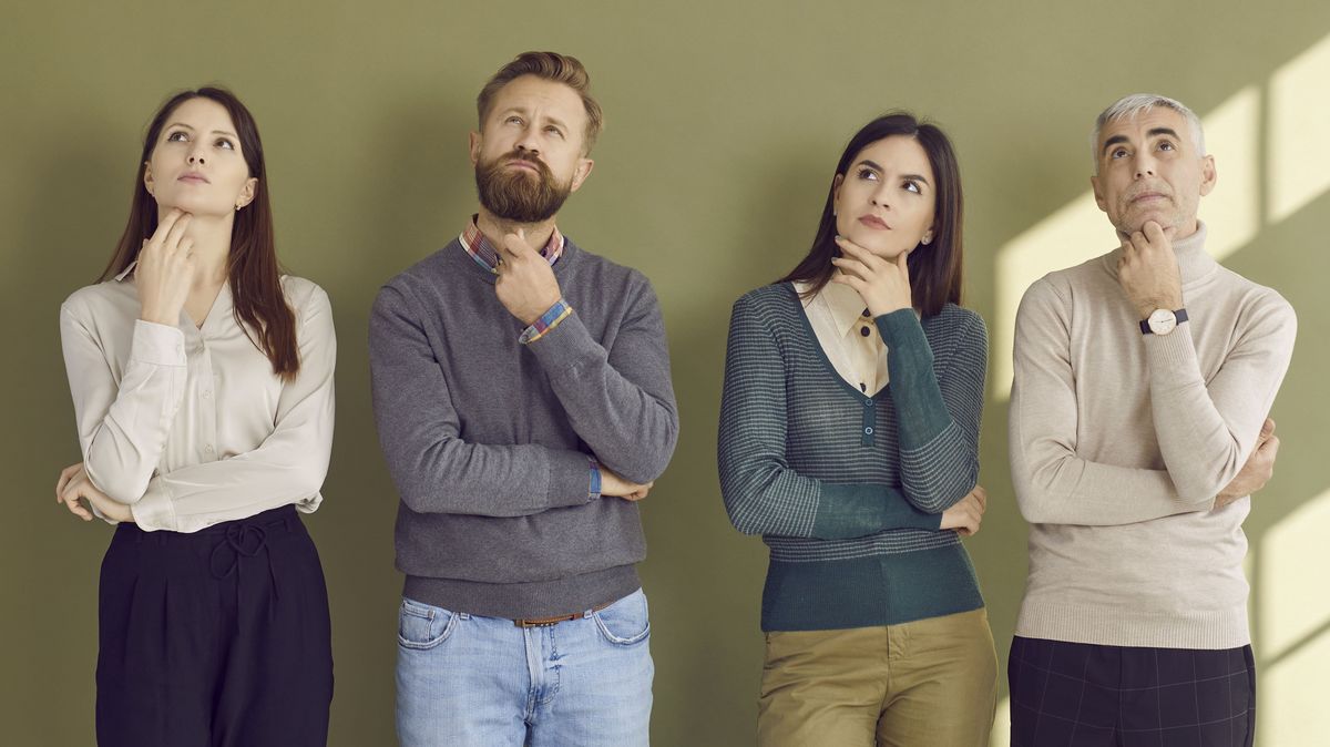 Farbfoto: Vier Menschen in formeller Kleidung stehen in einer Reihe nebeneinander und haben jeweils eine Hand ans Kinn gelegt. Sie wirken nachdenklich.