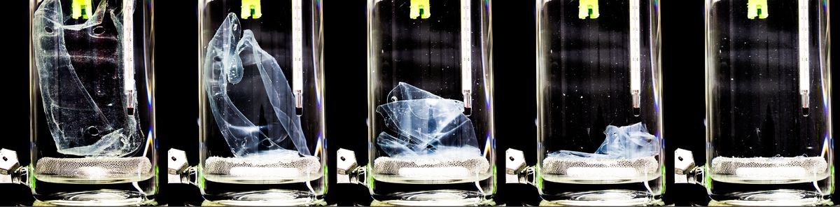 enlarge the image: Zu sehen sind nebeneinander fünf Gläser, die mit einer Flüssigkeit gefüllt sind. Im linken Glas ist ein größeres Plastikstück, im ganz rechten ist das Plastikstück komplett in der Flüssigkeit aufgelöst. Die Gläser dazwischen zeigen den Zerfallsprozess über einen Zeitraum von mehreren Stunden.
