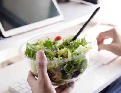 Eine Hand hält einen Salat, im Hintergrund ist ein Laptop zu sehen