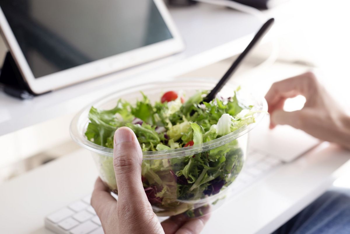 zur Vergrößerungsansicht des Bildes: Eine Hand hält einen Salat, im Hintergrund ist ein Laptop zu sehen
