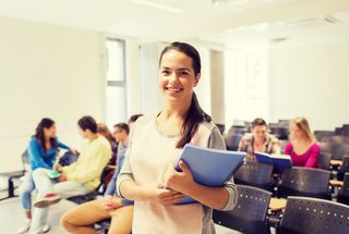 Eine junge Studentin steht lächelnd frontal vor der Kamera. Sie hält einen Schreibblock im Arm. Im Hintergrund sind weitere Studierende und leere Stühle zu sehen.