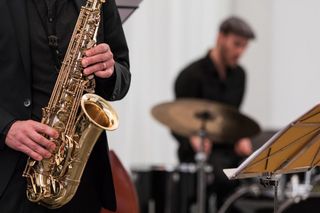 Das Foto bildet eine musikalische Szene ab. Links im Bild hält ein Musiker ein Saxophon, sein Kopf ist nicht zu sehen. Die rechte Bildhälfte zeigt unscharf die Umrusse eines weiteren Musikers, der Schlagzeug spielt, sowie den Notenständer des Saxophonisten.