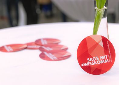 Eine Vase mit roter Tulpe auf einem weißen Stehtisch, an der Vase lehnt ein roter Sticker mit dem Claim "Sags mit Wisskomm"