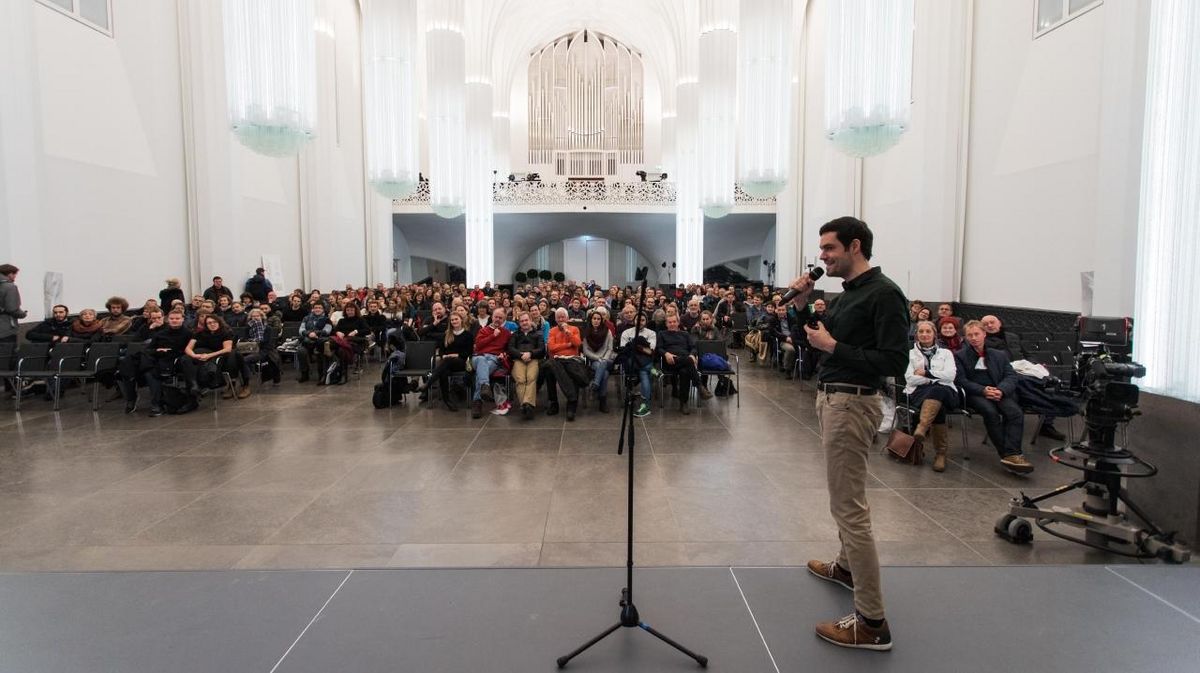 zur Vergrößerungsansicht des Bildes: Ein Mann steht auf einer Bühne im Paulinum. Er hält ein Mikrofon in der Hand. Im Hintergrund sitzt das Publikum.