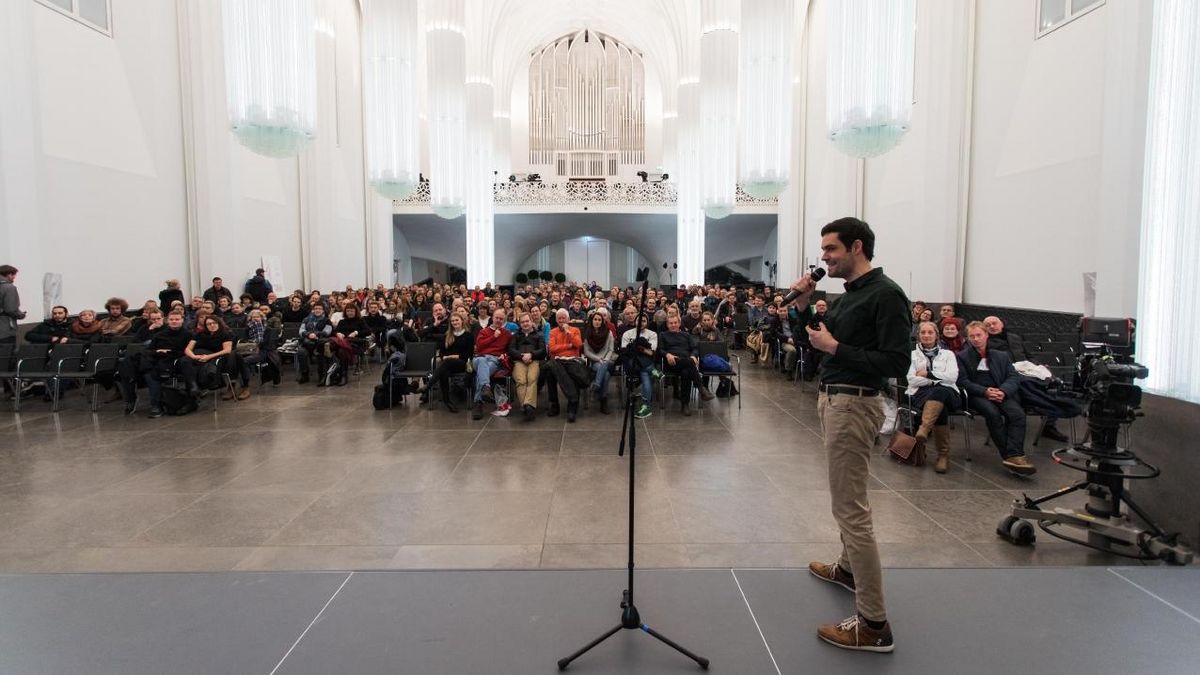 enlarge the image: Ein Mann steht auf einer Bühne im Paulinum. Er hält ein Mikrofon in der Hand. Im Hintergrund sitzt das Publikum.