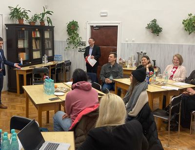 Ein Mann in einem dunkelblauen Anzug steht vorne in einem Seminarraum und spricht zu einer Gruppe von Teilnehmenden, die an Holztischen sitzen. IM Hintergrund befinden sich Bücherregale, Grünpflanzen und eine weiße Tafel.