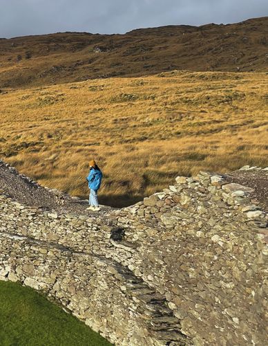 Eine Person läuft über eine breite Steinmauer. Im Hintergrund ist eine gelbe Wiese zu sehen, die eher moorartig ausieht.