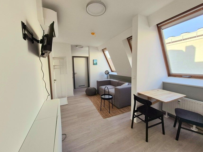 enlarge the image: Bright, minimalist room with a small couch, a wall-mounted TV, a table with two chairs, and large windows, featuring light wooden flooring and white walls. Photo: Thomas Biermann