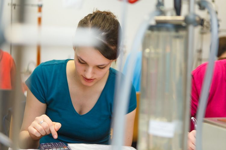 Studentin sitzt vor einem Versuchsaufbau und tippt etwas in ihren Taschenrechner ein, Foto: Christian Hüller
