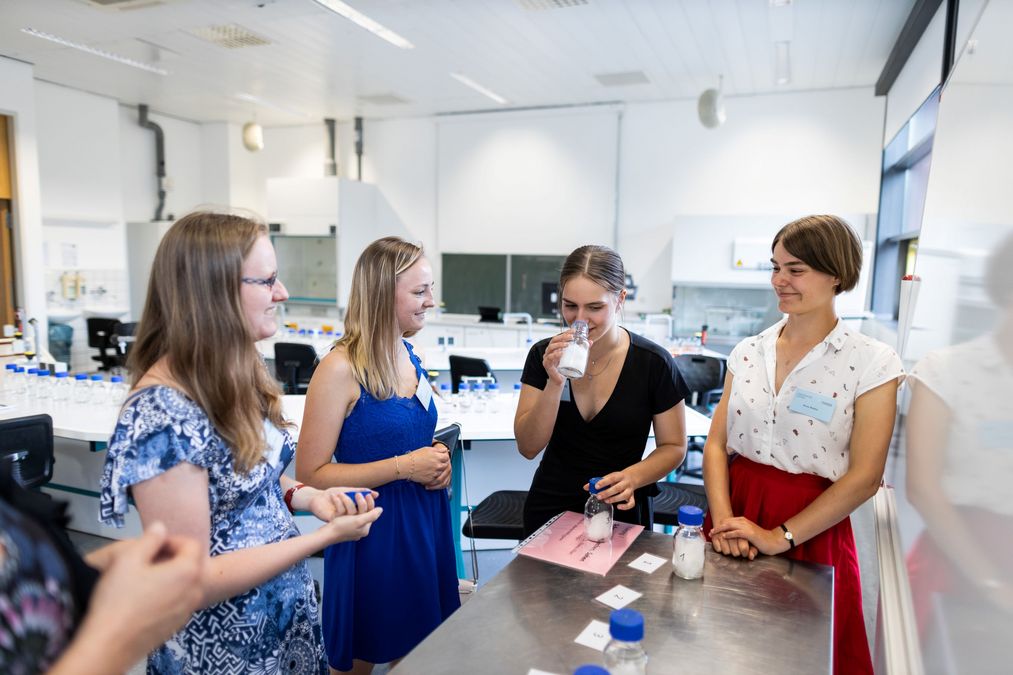 zur Vergrößerungsansicht des Bildes: Farbfoto: 4 Stipendiatinnen stehen an einem Tisch, auf dem Tisch stehen Flaschen mit weißem Inhalt, eine Stipendiatin riecht an einer Flasche