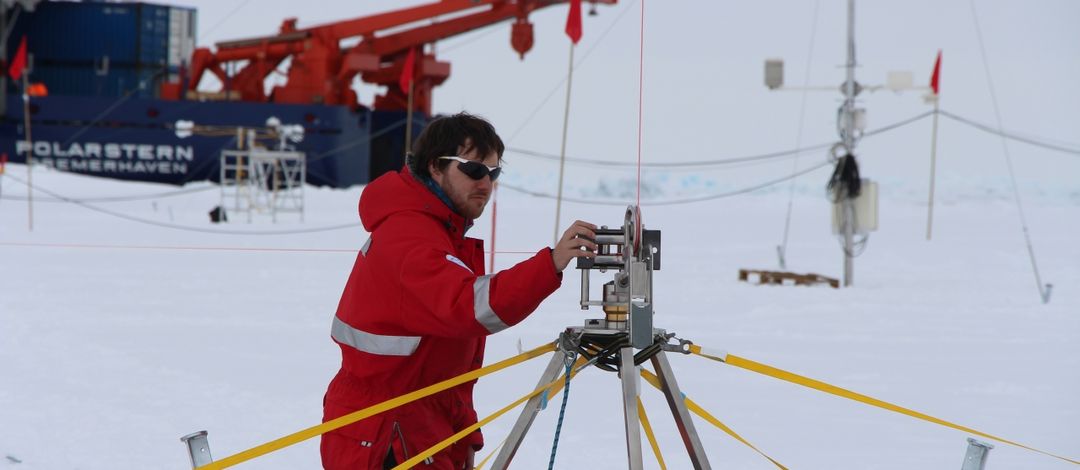 Ein Forscher baut eine Messstation im arktischen Eis auf. Im Hintergrund steht der Eisbrecher Polarstern.