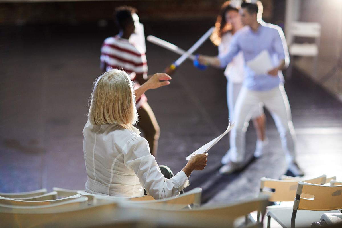 Das Bild zeigt eine Frau, die Regieanweisungen bei einer Theaterprobe gibt. Im Hintergrund sieht man Schauspieler, die einen Schwertkampf proben. 