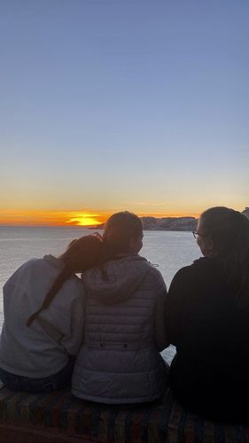 Drei Personen stehen mit dem Rücken zur Kamera und schauen auf das Meer hinaus. Die Sonne geht unter. Das Bild ist dadurch in dunklem Blau gehalten.