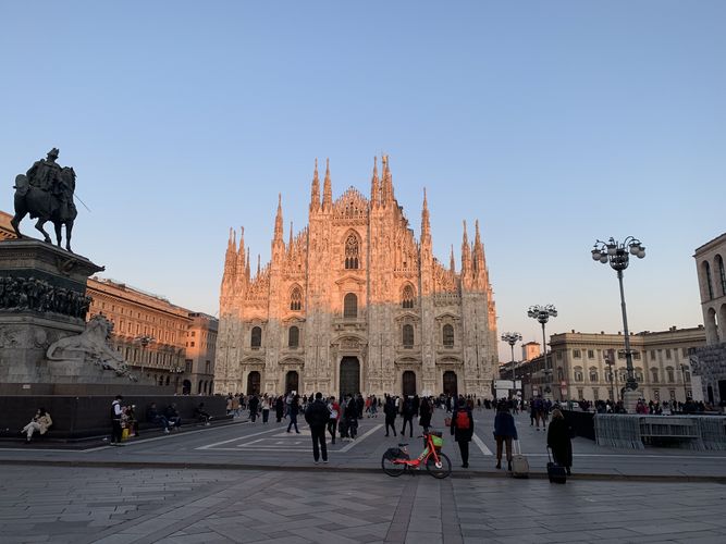 Farbfoto: Gebäudeaufnahme des Doms mit Türmen, Verzierungen und Figuren. Davor befindet sich ein Platz mit Menschen und einer Reiterstatue